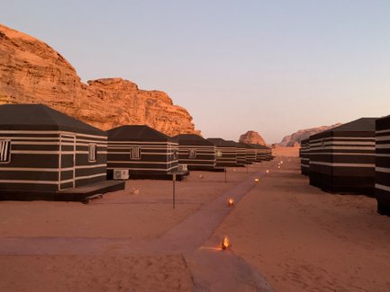 In der Wüste stehen mehrere schwarze Zelte mit weißen Streifen, umgeben von beeindruckenden Felsformationen im Abendlicht.