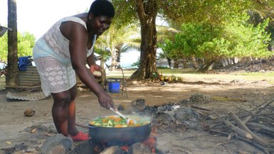 Eine Frau kocht im Freien in einem großen Topf über offenem Feuer, umgeben von Bäumen und einer entspannten Atmosphäre.