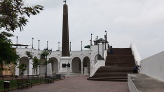 Ein monumentaler Obelisk mit einer Statue auf der Spitze steht in einem historischen Platz, umgeben von Bänken und Bäumen.