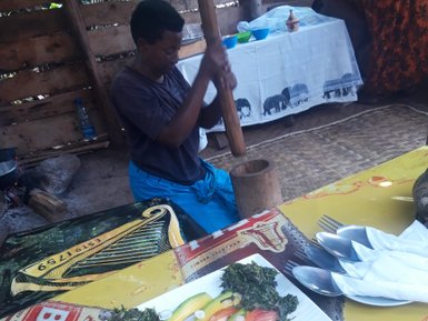 Eine Person stampft mit einem Holzstößel in einem traditionellen Mörser, während frisches Gemüse auf einem Teller präsentiert wird.