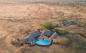 Eine Luftaufnahme eines luxuriösen Safari-Camps mit einem Pool, umgeben von trockener Savanne und sanften Hügeln im Hintergrund.