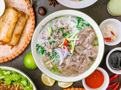 Eine Schüssel mit Pho, gefüllt mit zartem Rindfleisch, frischen Kräutern und roten Chilischoten, umgeben von Limetten.