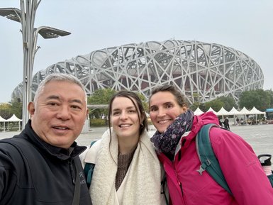 Drei Personen posieren fröhlich vor dem Olympiastadion in Peking, umgeben von einer modernen Architektur und grünen Bäumen.
