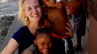 Eine lächelnde Frau umarmt zwei Kinder, während weitere Kinder im Hintergrund spielen und eine freundliche Atmosphäre schaffen.