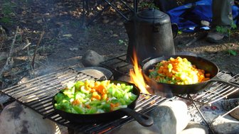 Zwei Pfannen mit buntem Gemüse, darunter Karotten und Sellerie, brutzeln über einem Lagerfeuer in der Natur.