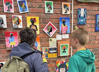 Zwei Personen betrachten eine Wand mit bunten Kunstwerken, die stilisierte Porträts schwarzer Frauen zeigen.