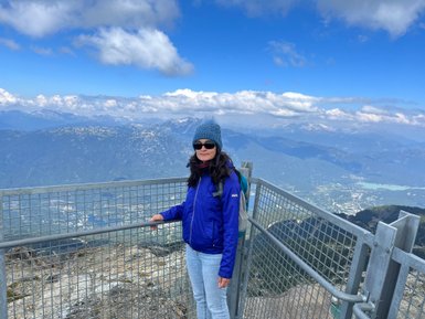 Eine Frau steht an einem Aussichtspunkt auf einem Berg, umgeben von beeindruckenden Wolken und Bergen im Hintergrund.