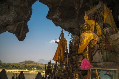 Im Inneren einer Höhle stehen zahlreiche goldene Buddha-Statuen, umgeben von bunten Blumen und religiösen Symbolen.