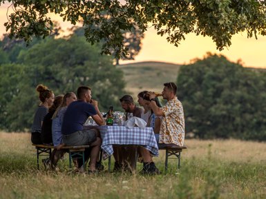 Eine Gruppe von Freunden sitzt an einem Tisch im Freien, umgeben von Natur, und genießt ein gemeinsames Essen bei Sonnenuntergang.