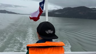 Ein Kind in einer orangefarbenen Schwimmweste schaut auf das Wasser, während eine Flagge im Wind weht.
