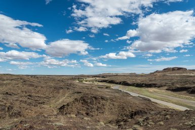 Eine weite, karge Landschaft mit sanften Hügeln und einem ruhigen Fluss, unter einem strahlend blauen Himmel mit Wolken.