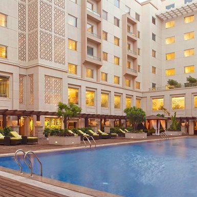 Ein eleganter Hotelpool mit Liegen und Palmen, umgeben von einem modernen Gebäude mit kunstvollen Fenstern.