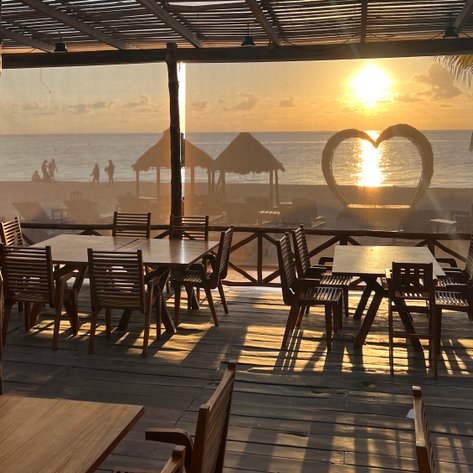 Ein wunderschöner Sonnenuntergang über dem Meer, sichtbar durch eine offene Restaurantterrasse mit Holztischen.