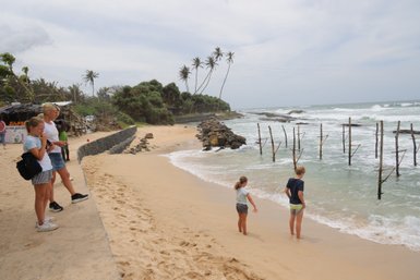 Am Strand stehen zwei Kinder und eine Frau, während die Wellen sanft an den Sand schlagen und Palmen im Hintergrund wehen.