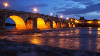 Eine beleuchtete Brücke reflektiert im Wasser, während der Himmel in sanften Blautönen leuchtet und die Stadt im Hintergrund sichtbar ist.
