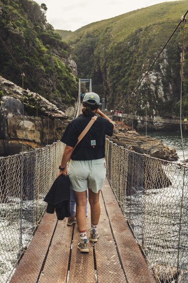 Eine Frau mit einem Rucksack überquert eine Hängebrücke, umgeben von üppiger Natur und steilen Klippen.
