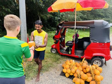 Ein Verkäufer überreicht einem Jungen eine frische Kokosnuss, während ein Tuktuk im Hintergrund parkt.