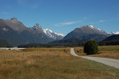 Eine kurvenreiche Schotterstraße führt durch eine weite, grüne Landschaft mit majestätischen Bergen im Hintergrund.