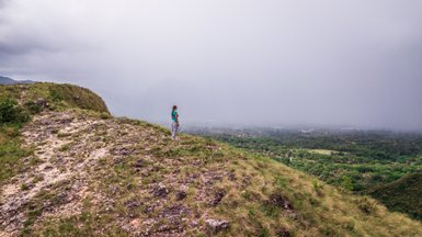 Eine Frau im blauen Oberteil steht auf einem Hügel und blickt nachdenklich auf die bewaldete Landschaft in der Ferne.