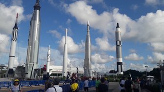 Eine Gruppe von Besuchern steht vor mehreren beeindruckenden Raketen im Kennedy Space Center unter einem blauen Himmel.