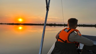 Ein Junge in einer orangefarbenen Schwimmweste sitzt in einem Boot und beobachtet den Sonnenuntergang über einem ruhigen Gewässer.