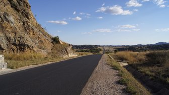 Eine asphaltierte Straße schlängelt sich durch eine weite, trockene Landschaft mit sanften Hügeln und Felsen im Hintergrund.