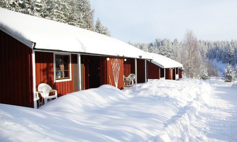 Rote Hütten stehen in einer schneebedeckten Landschaft, umgeben von hohen, verschneiten Bäumen und klarem Himmel.