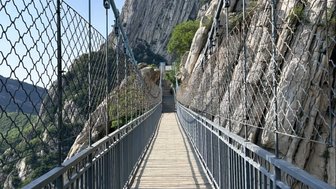 Eine schmale Hängebrücke führt über eine Schlucht, umgeben von steilen Felsen und üppigem Grün im Hintergrund.