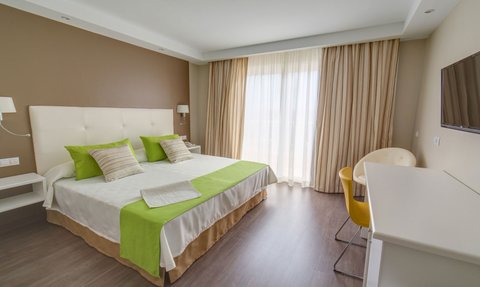 Ein helles Hotelzimmer mit zwei Betten, grünen Kissen und einem großen Fenster, das Tageslicht hereinlässt.