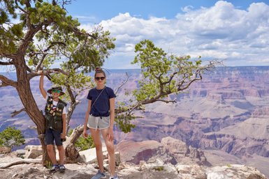 Ein Junge mit Sonnenbrille und Hut steht an einem Baum, während eine Frau in sportlicher Kleidung lächelt, mit dem Grand Canyon im Hintergrund.