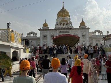 Zwei Personen mit orangefarbenen Turbanen stehen vor dem Gurudwara Sri Bangla Sahib, umgeben von einer lebhaften Menschenmenge.