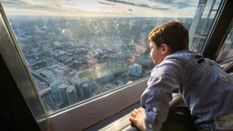 Ein Junge schaut fasziniert aus einem hohen Fenster auf die Stadt Toronto, während die Sonne am Horizont untergeht.