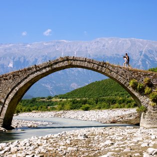 Ein Paar geht über eine alte Steinbrücke, umgeben von grünen Hügeln und majestätischen Bergen unter einem strahlend blauen Himmel.