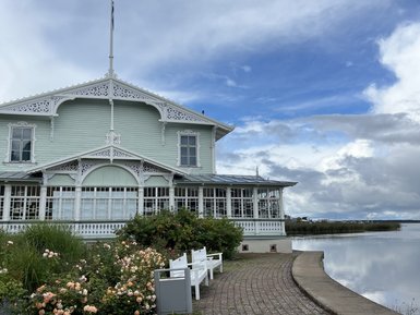 Ein elegantes, hellgrünes Gebäude mit verzierten Fenstern steht am Ufer eines ruhigen Gewässers, umgeben von blühenden Rosen.