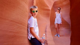 Ein Junge mit Sonnenbrille lehnt an einer roten Felswand und hält eine Wasserflasche, während ein Mädchen im Hintergrund steht.