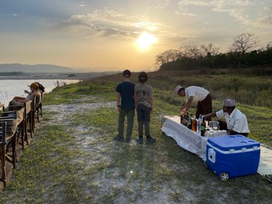 Zwei Kinder stehen am Ufer eines Flusses, während im Hintergrund ein Sonnenuntergang die Landschaft erleuchtet.
