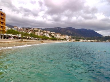 Ein ruhiger Strand mit klarem Wasser, umgeben von modernen Gebäuden und sanften Hügeln unter einem bewölkten Himmel.