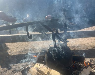Ein rauchender Lagerfeuerplatz mit einem Kessel, der über dem Feuer hängt, während Würstchen grillen.