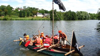 Eine Gruppe von Menschen in orangefarbenen Schwimmwesten paddelt in einem Boot auf einem ruhigen Fluss unter blauem Himmel.