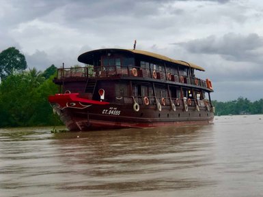 Ein traditionelles Holzboot mit rotem Bug schwebt sanft auf dem ruhigen Wasser, umgeben von üppigem Grün und bewölktem Himmel.