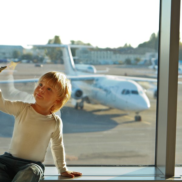 Ein kleiner Junge sitzt am Fenster eines Flughafens und hält ein Spielzeugflugzeug in der Hand, während er nach draußen schaut.
