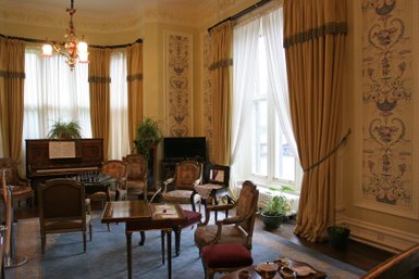 Ein elegantes Zimmer mit einem Klavier, umgeben von antiken Möbeln und großen Fenstern, die viel Tageslicht hereinlassen.