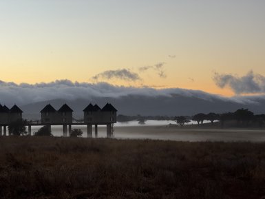 Die stilvollen Lodges stehen auf Stelzen in einer nebligen Landschaft, während die Sonne hinter den Bergen aufgeht.