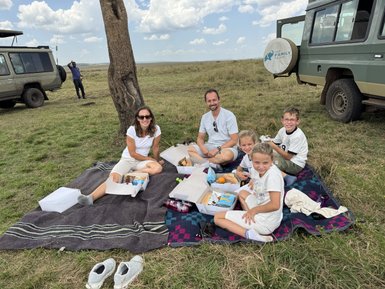 Eine Familie genießt ein Picknick auf einer Decke im Freien, umgeben von der weiten Landschaft der Masai Mara.