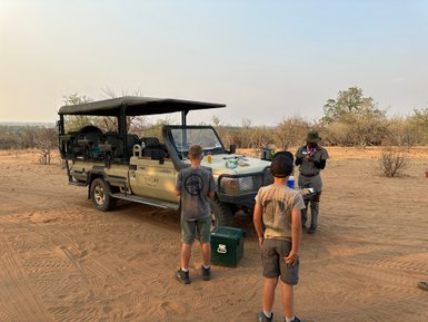 Ein Jeep steht in der Wüste, während zwei Kinder und ein Guide sich um einen Tisch mit Snacks versammeln.