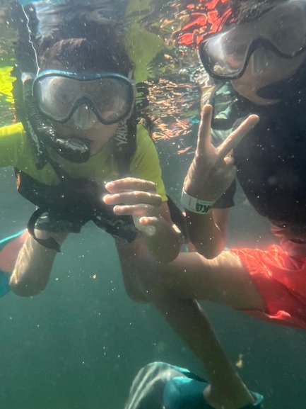 Zwei Kinder tauchen unter Wasser, tragen Schnorchel und zeigen ein Peace-Zeichen, umgeben von glitzerndem Wasser.
