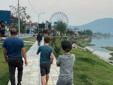 Eine Familie spaziert am Phewa-See entlang, während im Hintergrund ein Riesenrad zu sehen ist.