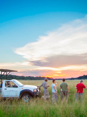 Eine Gruppe von fünf Personen steht in einem weiten Grasfeld, während die Sonne am Horizont untergeht.