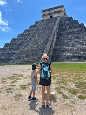 Eine Frau mit einem Rucksack und ein Junge stehen vor der beeindruckenden Pyramide von Chichen Itza an einem sonnigen Tag.