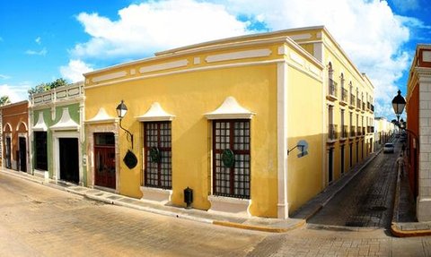 Eine sonnige Straßenszene mit bunten Gebäuden, darunter ein gelbes Haus mit großen Fenstern und grünen Pflanzen.